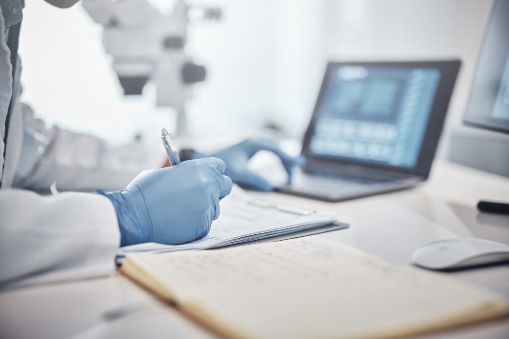 Scientist in white lab coat and blue rubber gloves writing down information from computer onto paper in a folder.
Picture by J Bettencourt.